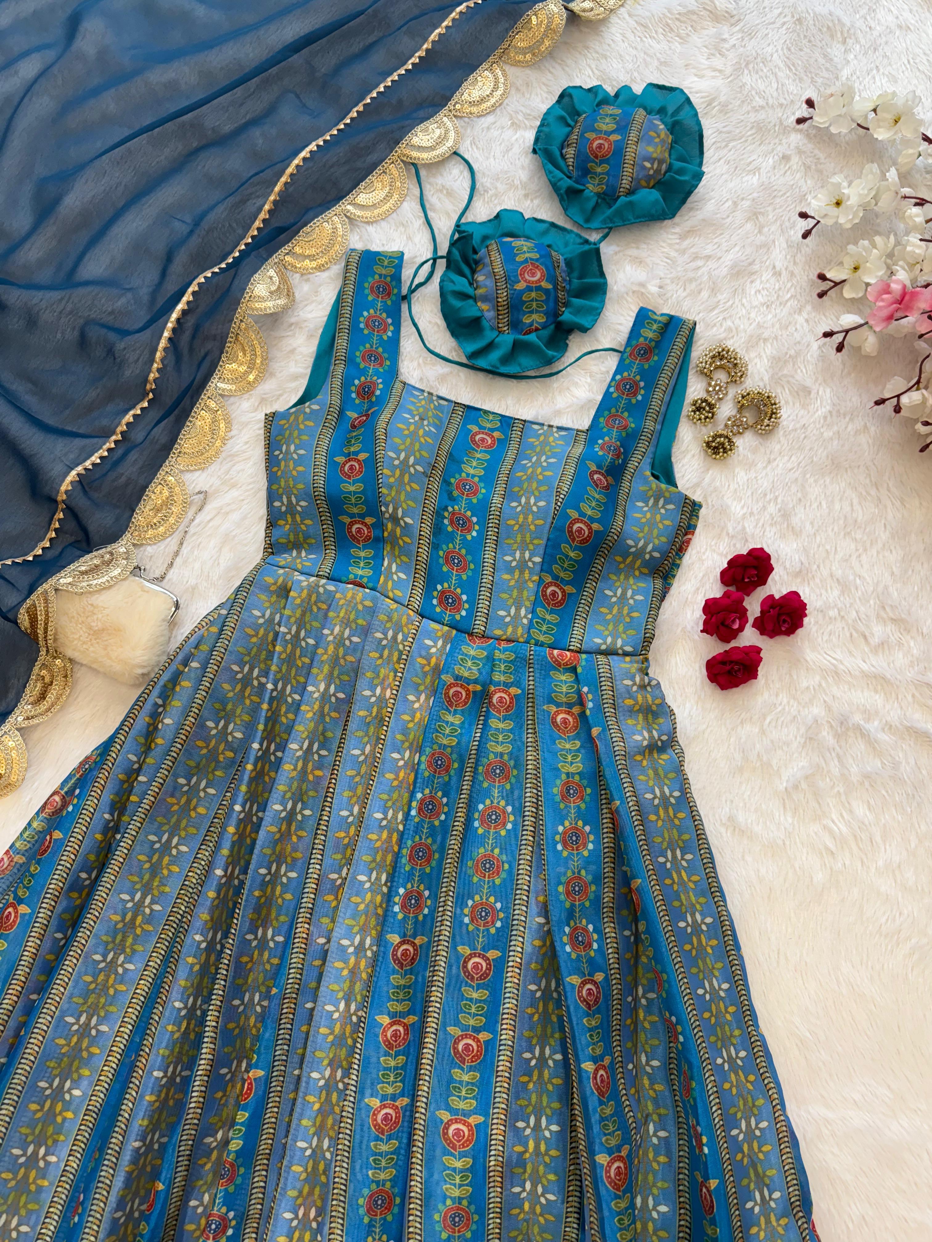 A blue patterned sleeveless dress laid flat on a white textured surface, styled with a matching blue dupatta, two blue flower-shaped accessories, red rose petals, and gold coin-like embellishments.
