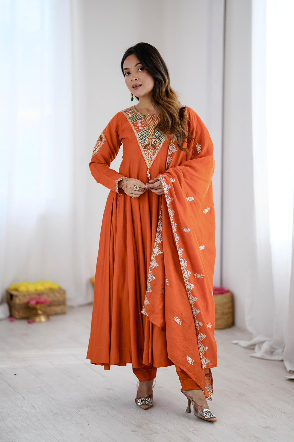 A woman models a traditional Indian outfit consisting of an orange kurta with gold embroidery, matching orange trousers, and an orange dupatta with white floral patterns, set against a bright, minimalist studio background with white curtains.