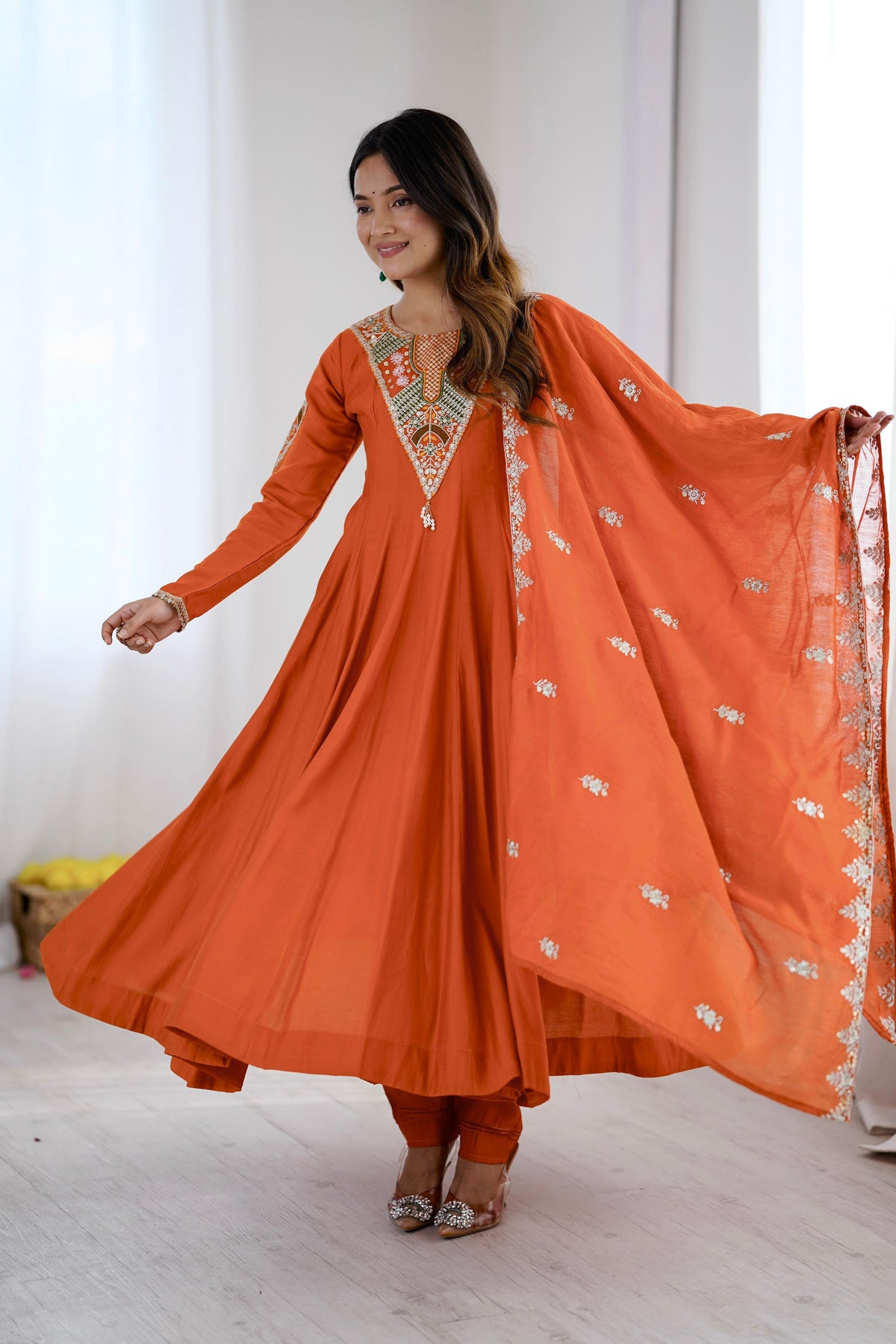 A woman modeling a vibrant orange traditional Indian outfit, featuring a long-sleeved kurta with intricate embroidery around the neckline, matching orange pants, and a sheer orange dupatta with silver motifs, set against a bright, minimalist indoor background.