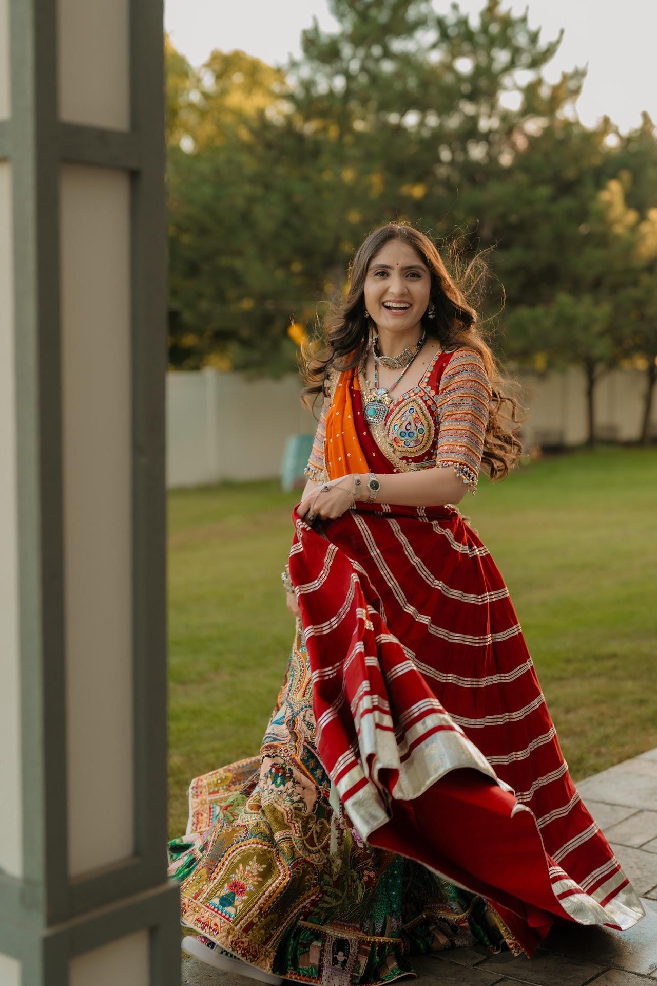 A woman wearing a traditional Indian red saree with white stripes and a heavily embroidered multicolor skirt, standing outdoors in a garden setting.