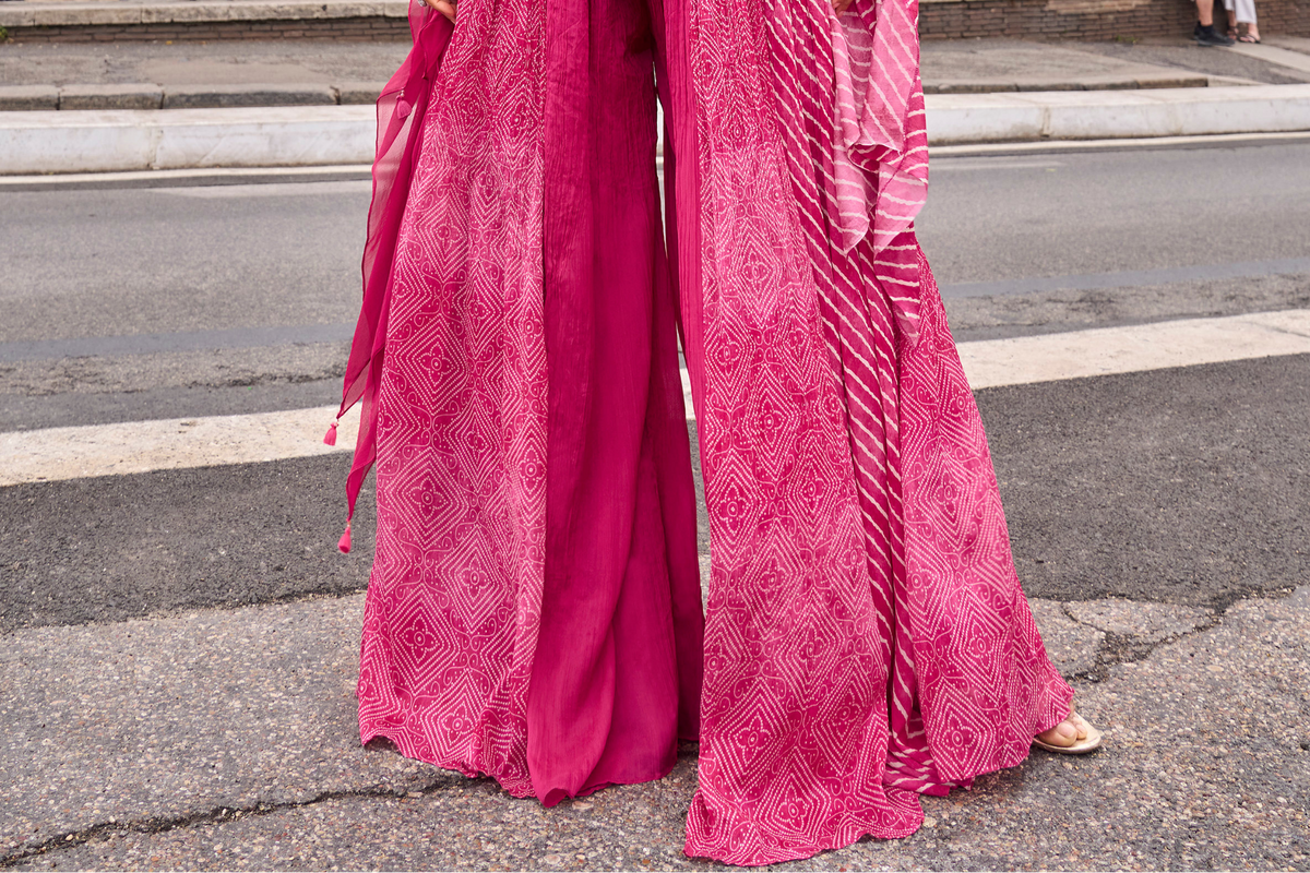 A close-up outdoor shot of two models wearing matching bright pink patterned maxi gharara pants with wide flared hems, standing on an asphalt street.